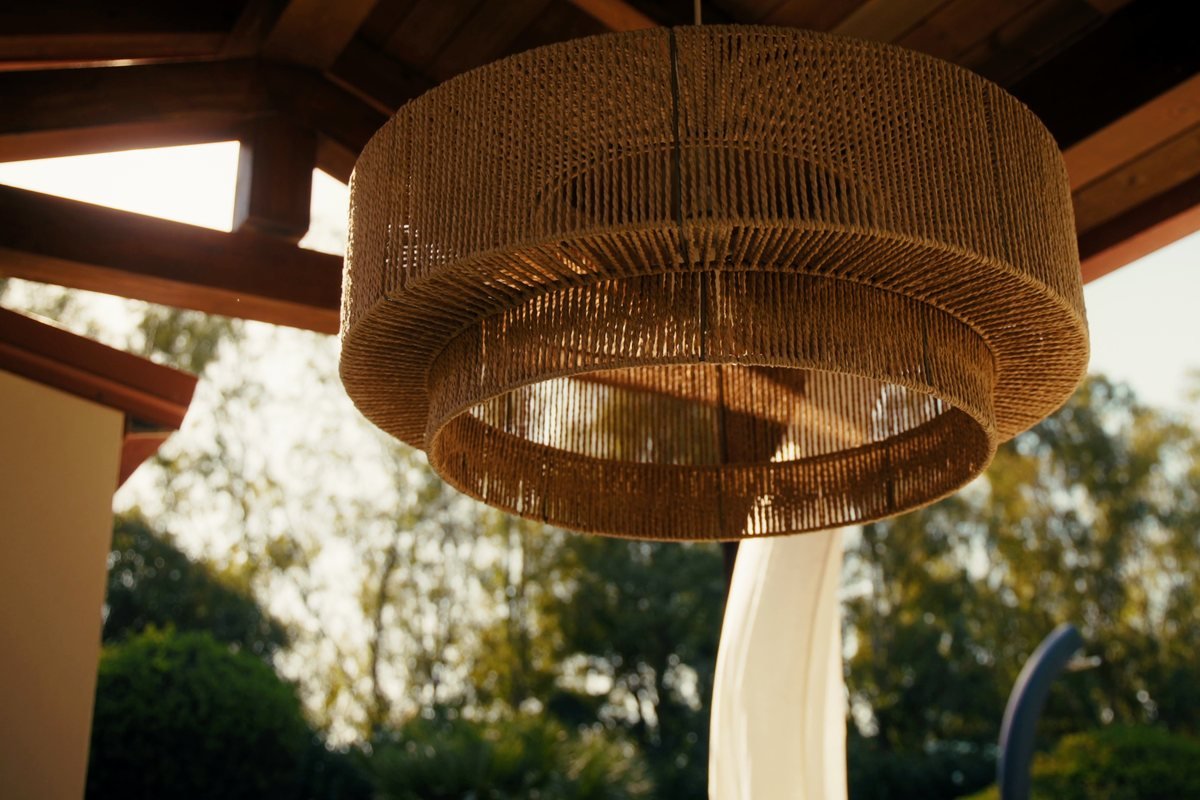 A woven lamp under a timber roof, the garden in sun behind it