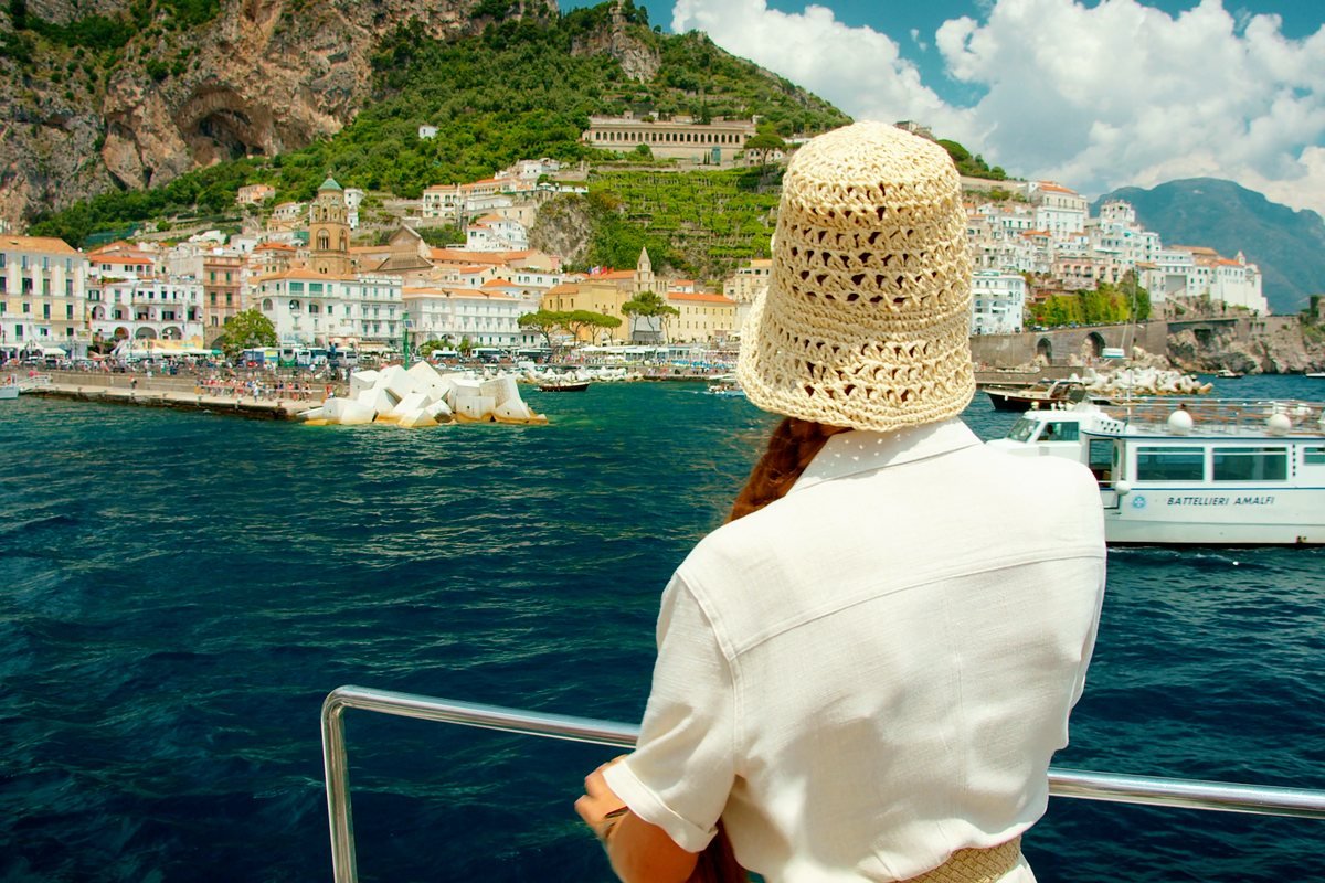 A woman in white linen and a straw hat on a boat coming into the Amalfi coast