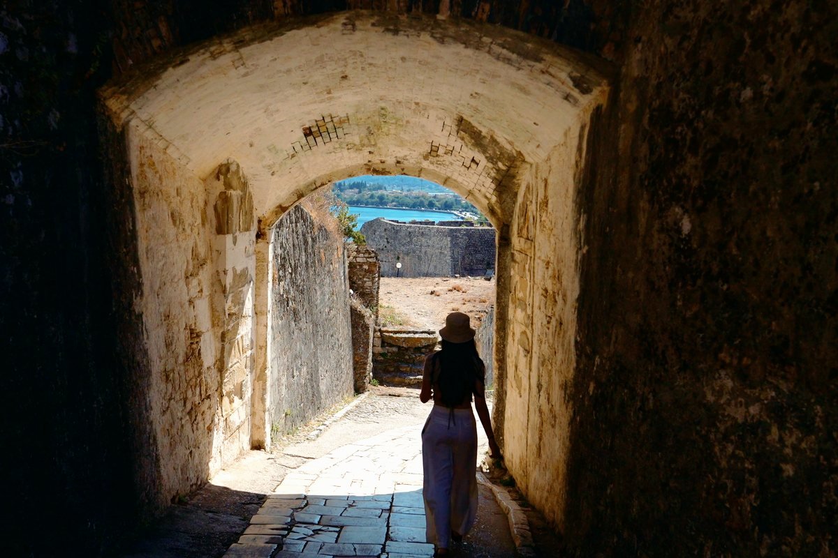 A woman walks out of a stone passage into sunlight above the sea