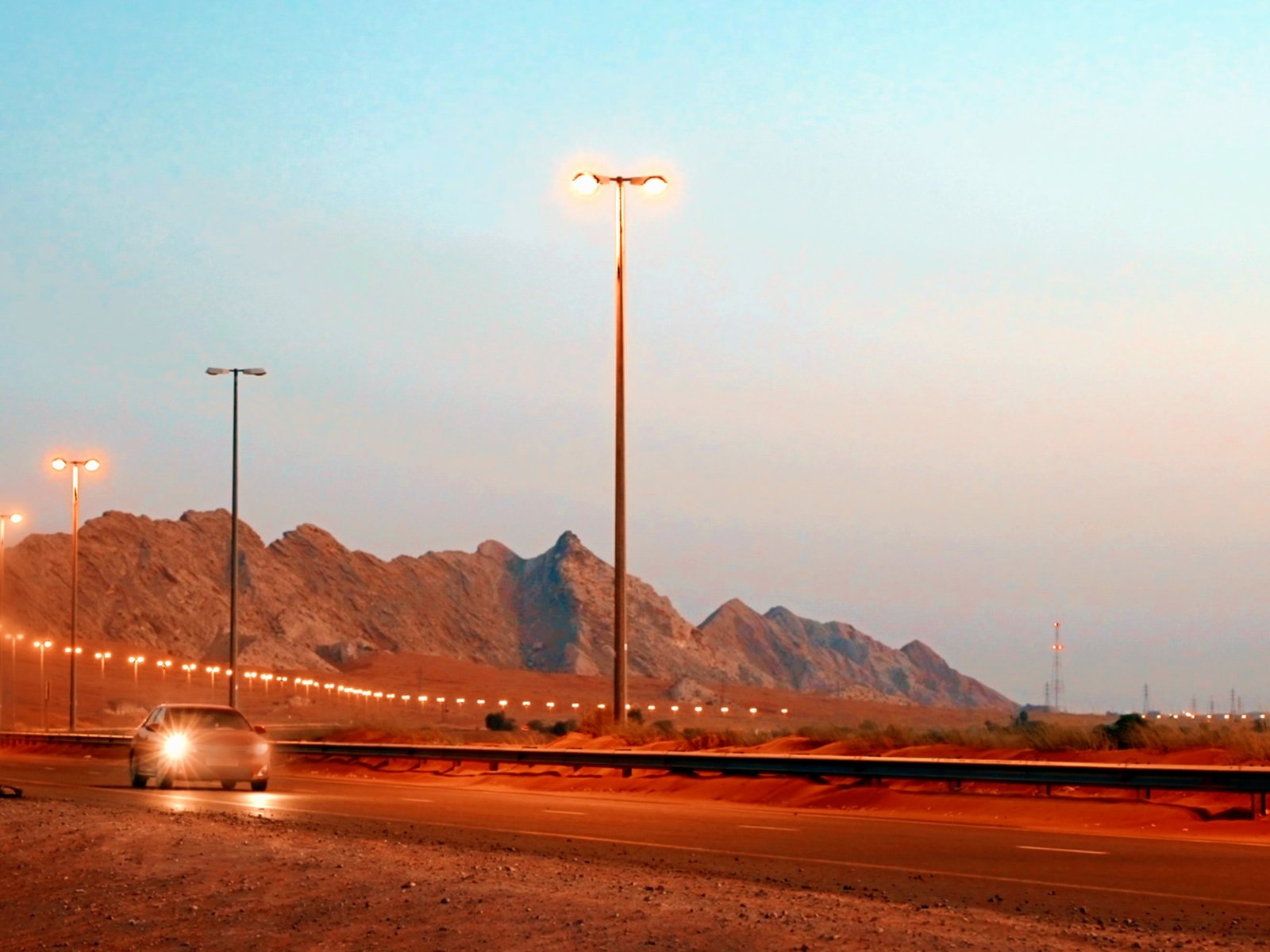 A desert highway at dusk below bare mountains, its lamps lit and a car approaching with its headlights on