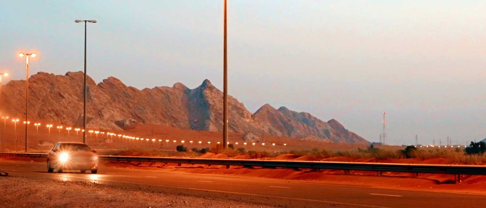 A desert highway at dusk below bare mountains, its lamps lit and a car approaching with its headlights on