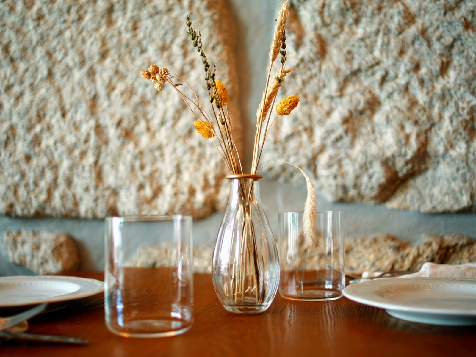 A table laid for two against a rough stone wall, dried wheat in a glass vase