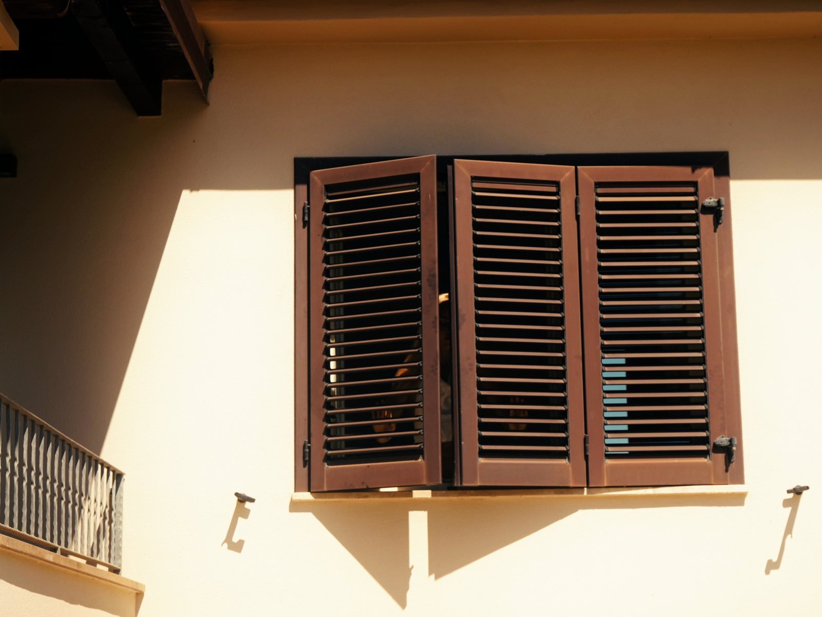 Wooden shutters being opened onto hard sun on a warm plaster wall