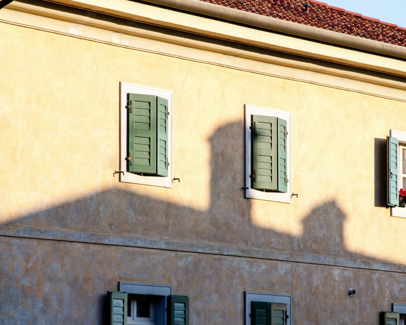 A plastered house front in low sun, green shutters, the shadow of the roofs across it